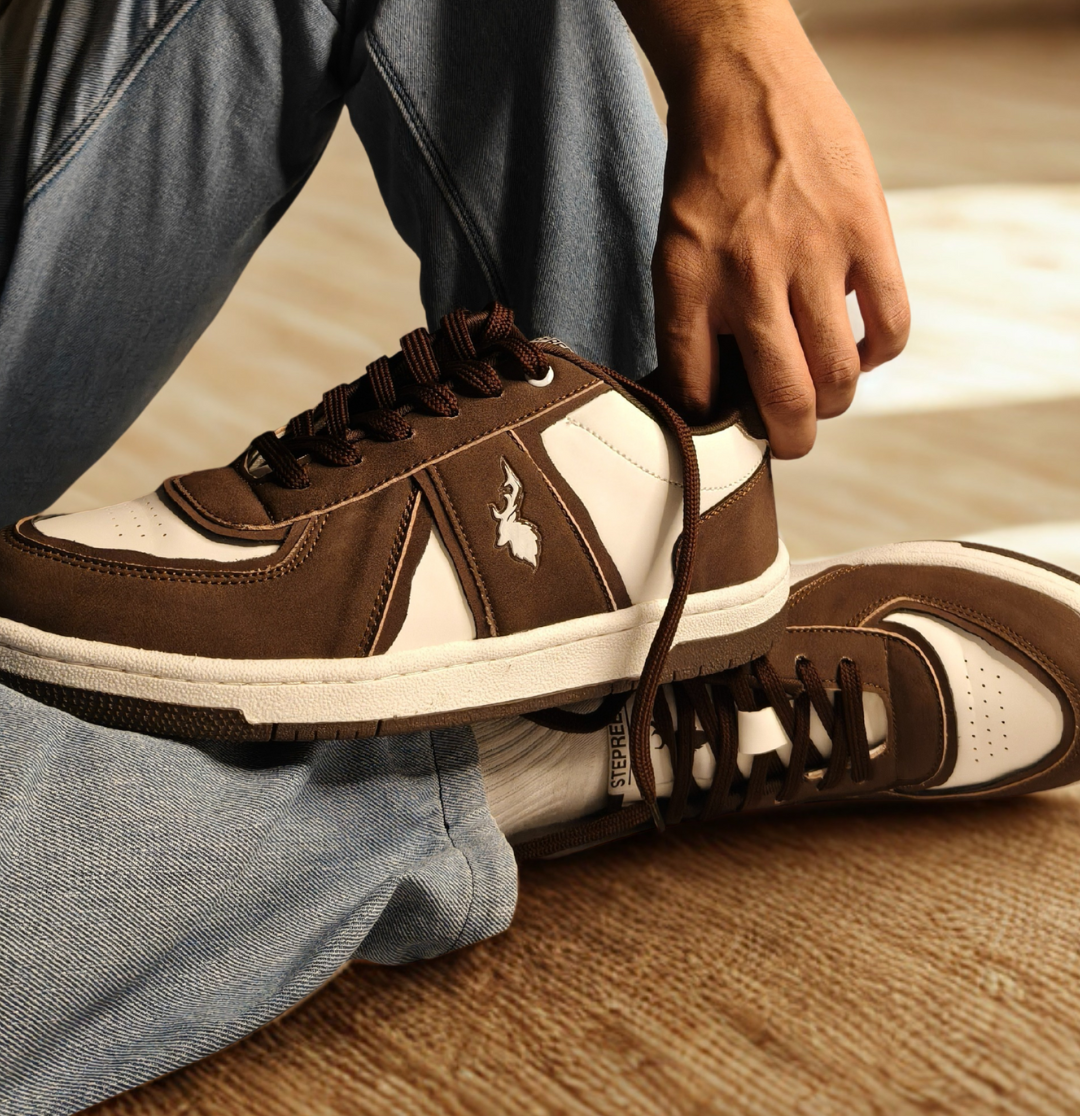 Brown and white casual sneakers with suede finish and contrast stitching, worn with blue jeans on a textured indoor floor, close-up view showing lace-up detail and branding