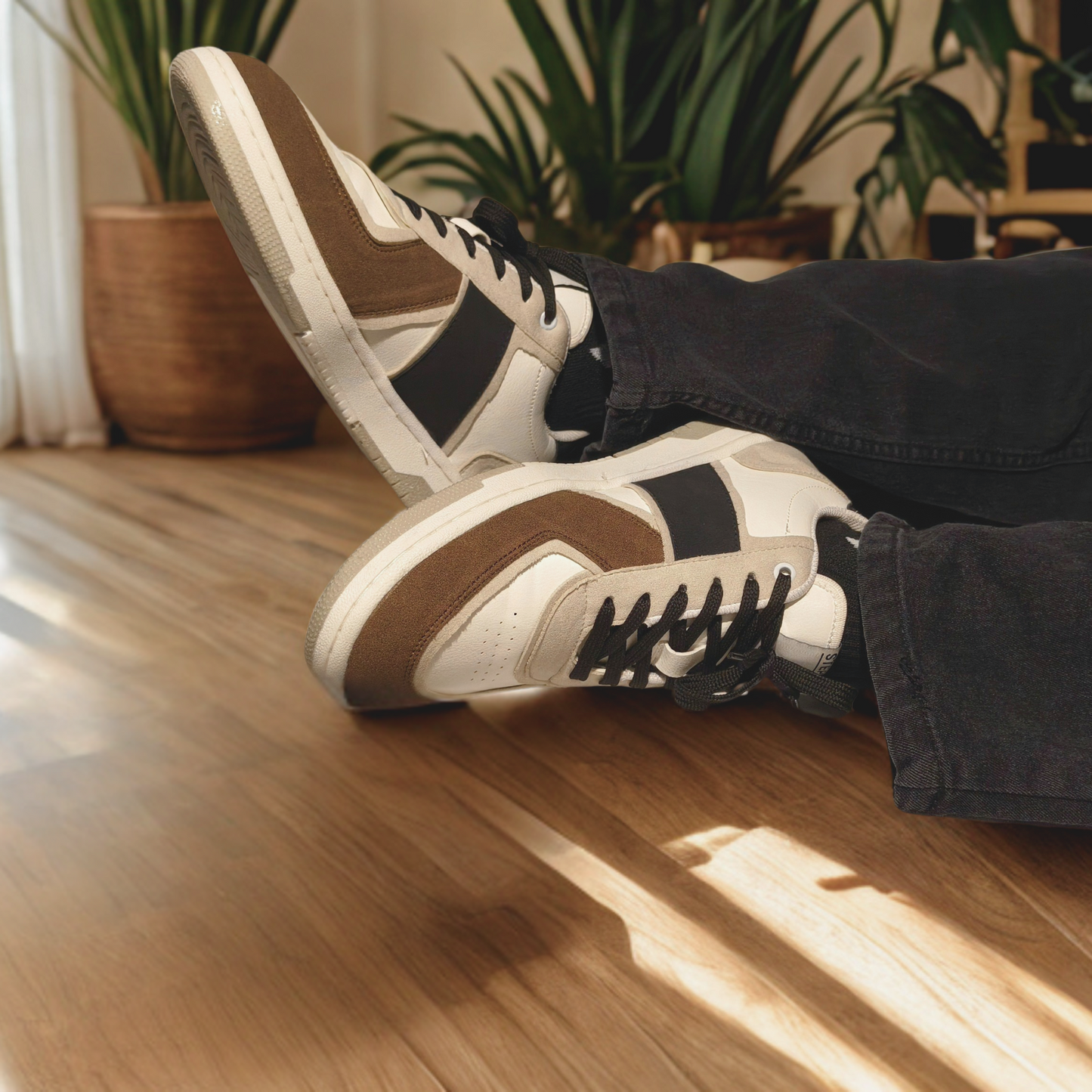 "On-feet view of brown, black, and white casual sneakers with suede overlays, worn with black jeans, styled indoors on wooden flooring with natural lighting and houseplants