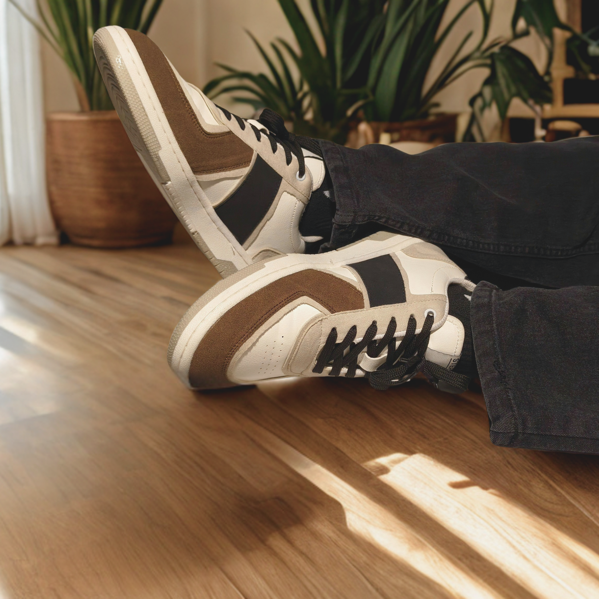 "On-feet view of brown, black, and white casual sneakers with suede overlays, worn with black jeans, styled indoors on wooden flooring with natural lighting and houseplants